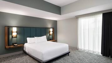 A hotel room with a large bed, a headboard with wall-mounted lights, a bedside table with a phone, and a window with sheer curtains.