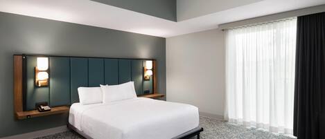A hotel room with a large bed, a headboard with wall-mounted lights, a bedside table with a phone, and a window with sheer curtains.