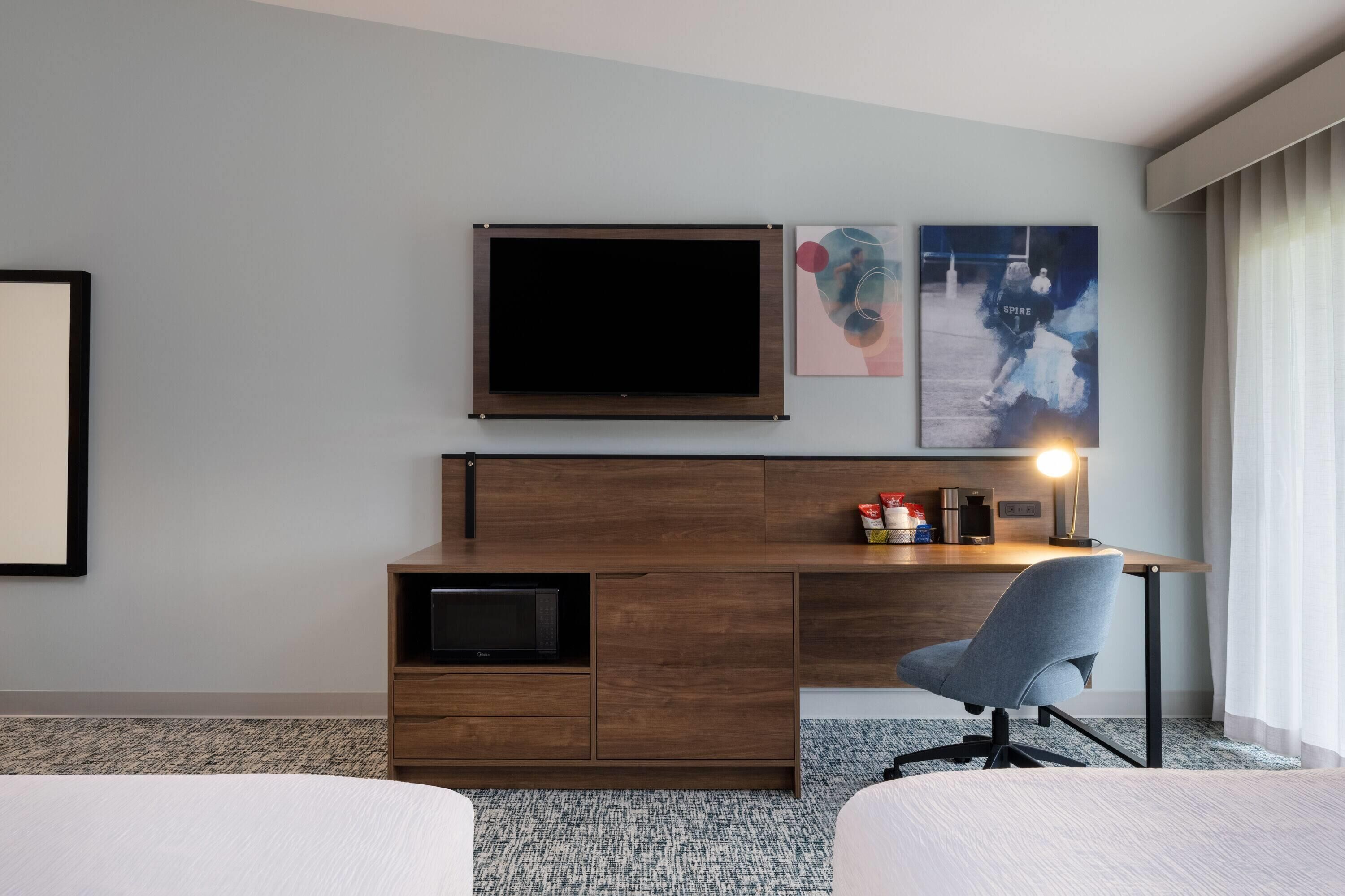 A hotel room with a bed, a desk with a chair, a TV, a coffee maker, and a framed picture on the wall.