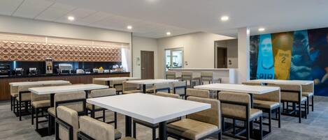 A modern dining area with multiple tables and chairs, a kitchen in the background, and a mural on the wall.