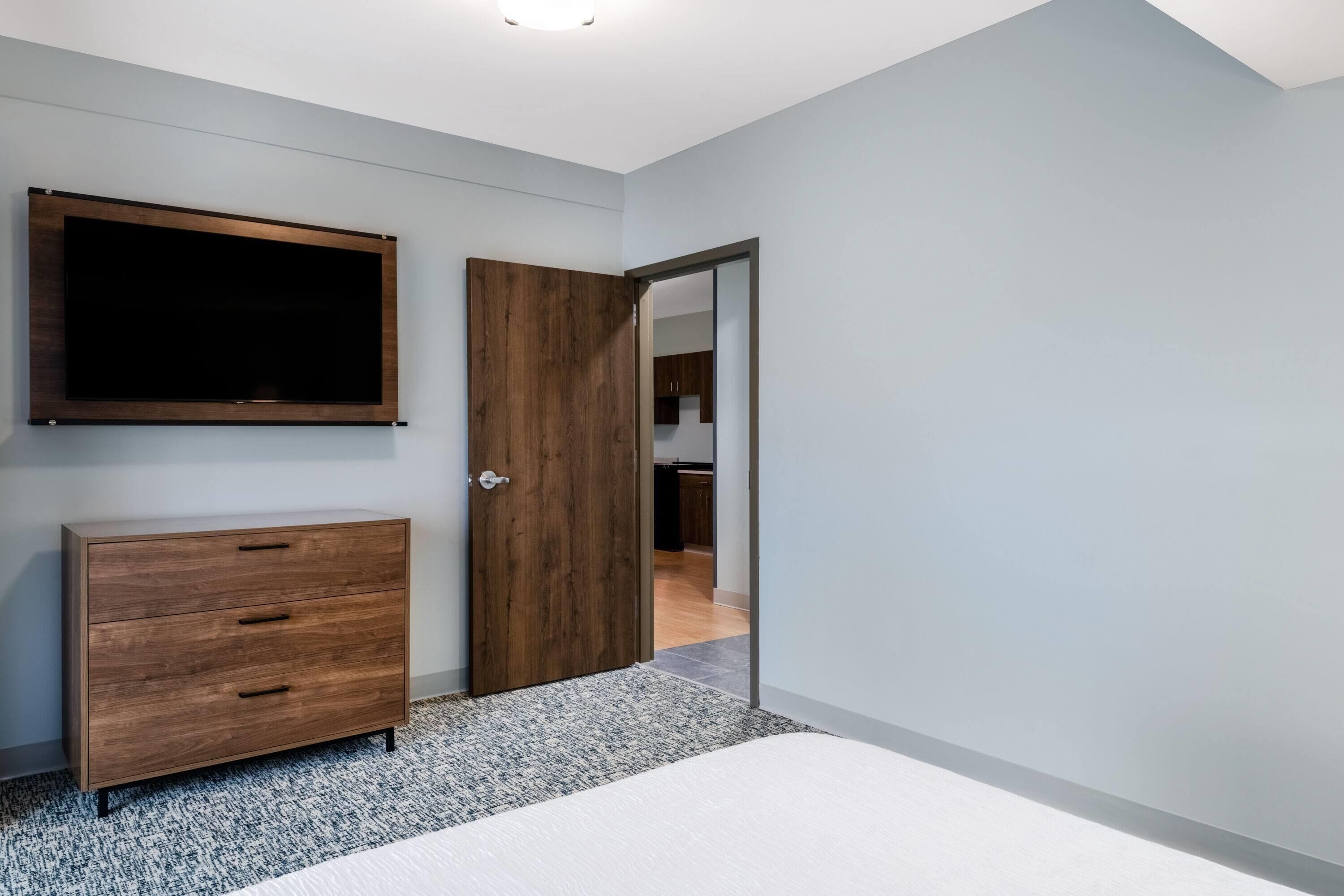 A bedroom with a bed, a wooden dresser, a TV mounted on the wall, and a door leading to another room.