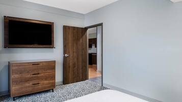 A bedroom with a bed, a wooden dresser, a TV mounted on the wall, and a door leading to another room.