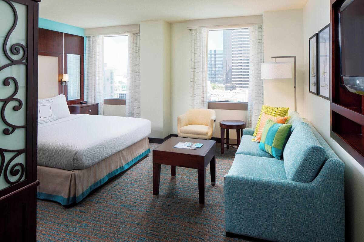 A hotel room with a large bed, a sofa, a small table, and a chair. There are windows with curtains, a lamp, and framed artwork on the wall.