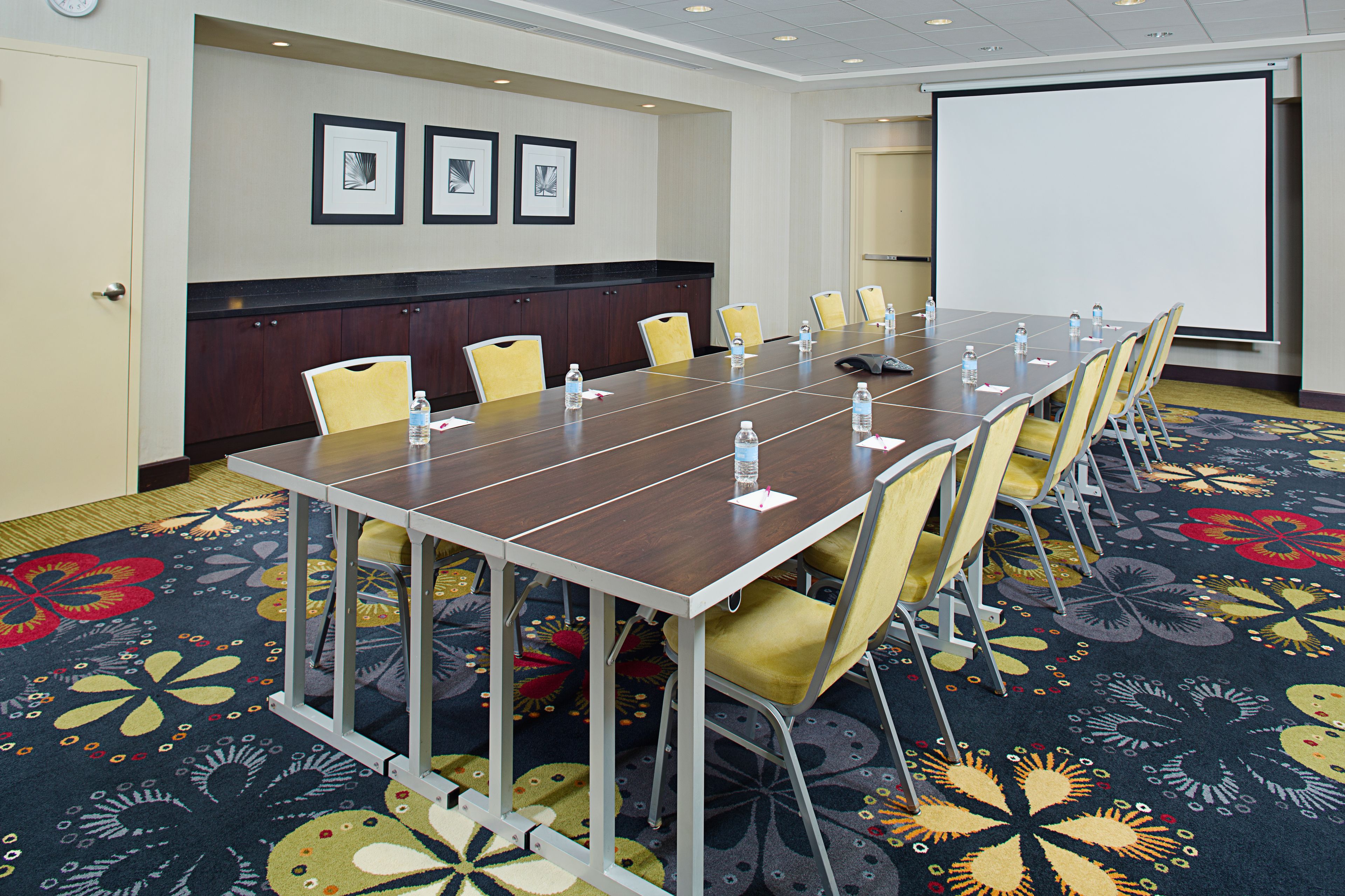 A conference room with a long table, chairs, a projector screen, and a carpet with floral patterns.