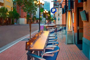 Un área de comedor al aire libre con una mesa larga y sillas, vista a la calle y un edificio con un letrero de estacionamiento 'P'.