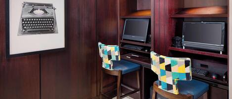 A workspace with two desks, each with a computer monitor and keyboard. There are two chairs with checkered covers. A framed picture of a typewriter is on the wall.