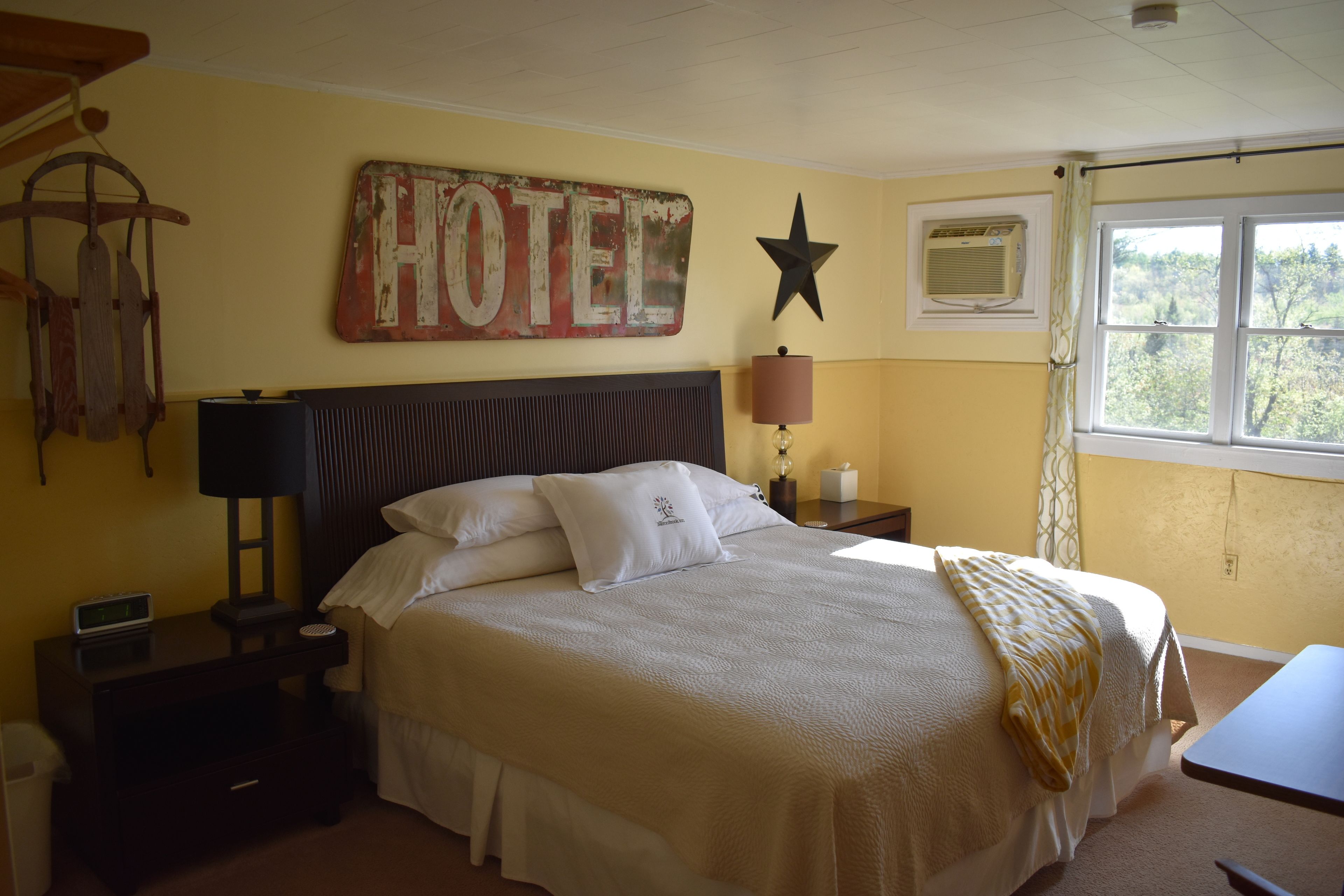 Ein Hotelzimmer mit Bett, Nachttisch, Lampe, Wanduhr, einer Stern-Dekoration und einem Fenster mit Blick auf Bäume.