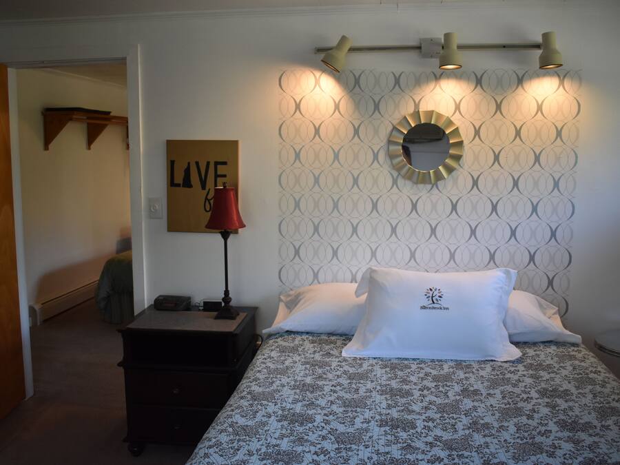 Un dormitorio ordenado con un papel tapiz estampado, una mesita de noche con lámpara, una cama con colcha floral y un espejo montado en la pared.
