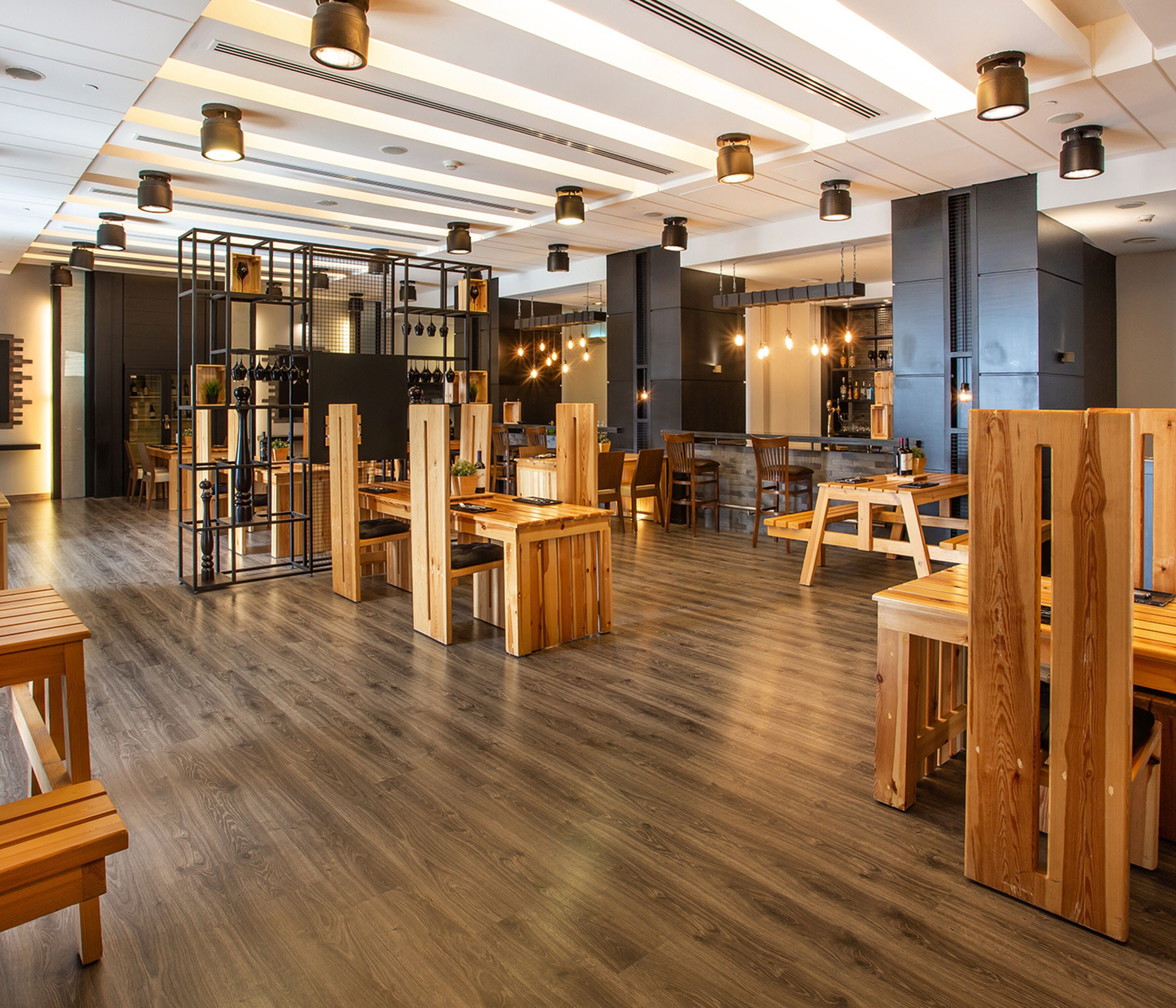 Interior moderno de restaurante con mesas y sillas de madera, zona de bar e iluminación colgante.