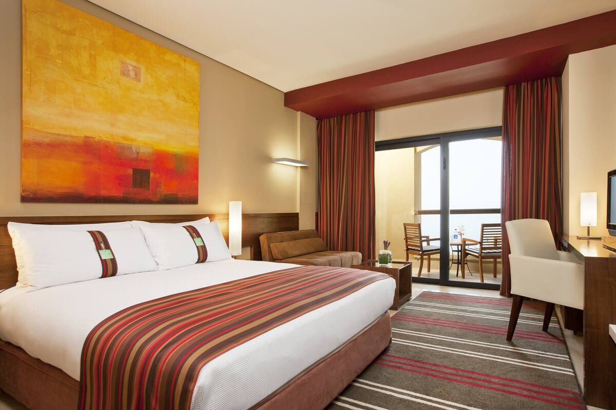 A hotel room with a large bed, a desk, a chair, a TV, and a view of the outside.