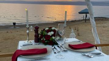 Una mesa dispuesta para dos personas, con velas, un ramo de flores y vista al mar al atardecer.