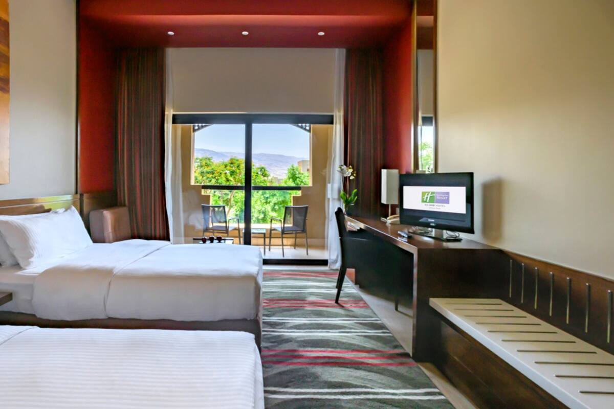 A hotel room with two beds, a desk with a TV, a phone, and a view of greenery and mountains.