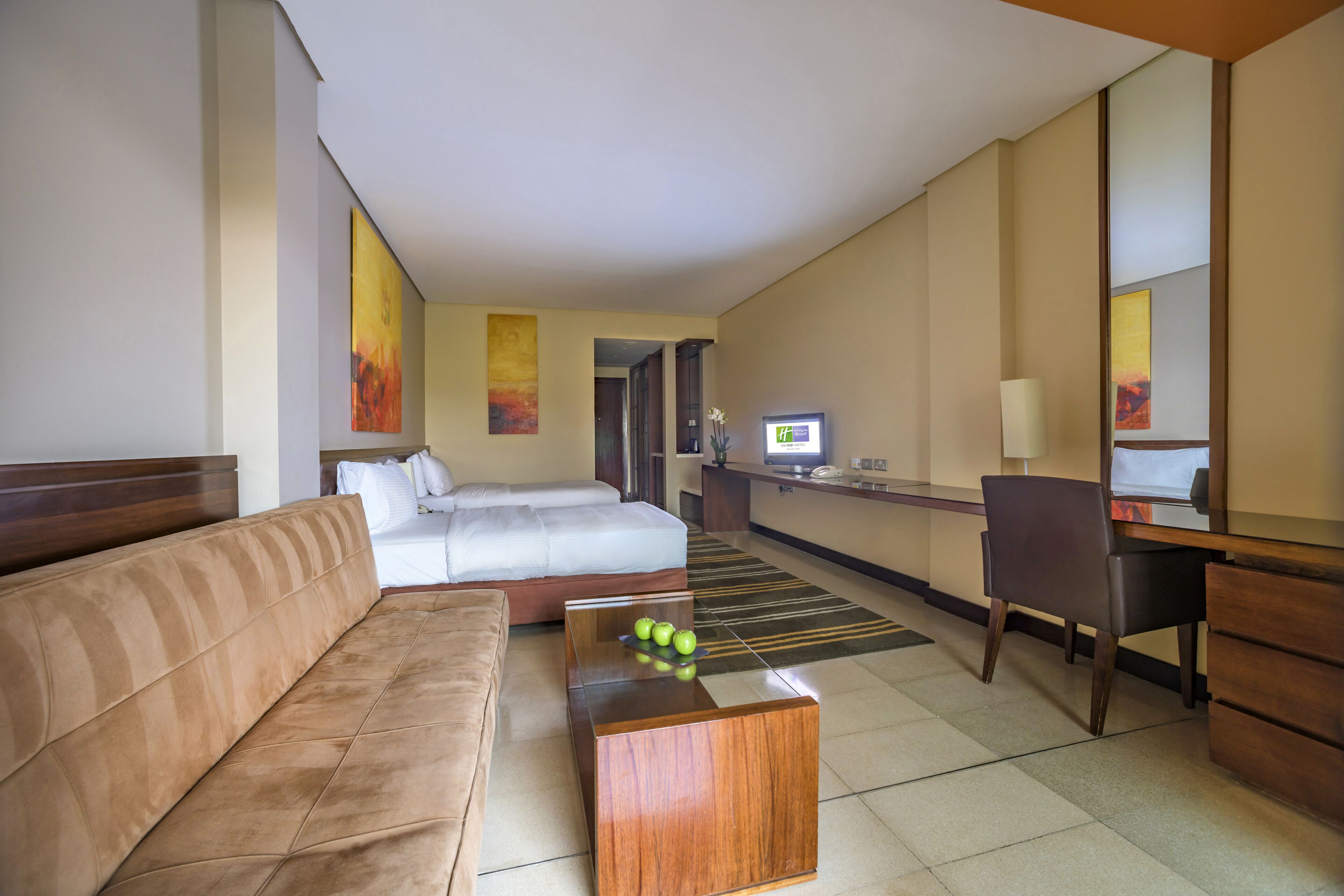 A hotel room with a sofa, a bed, a desk, a computer, and a small table with apples.