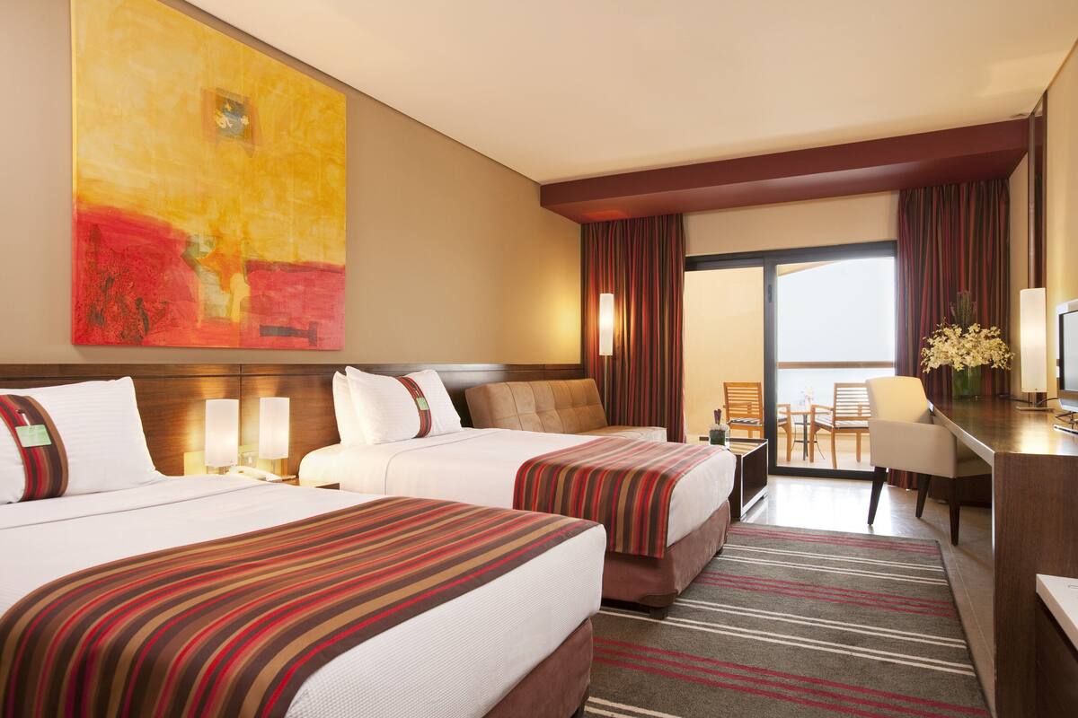 A hotel room with two beds, a large abstract painting, a desk with a chair, and a view of the outdoors.