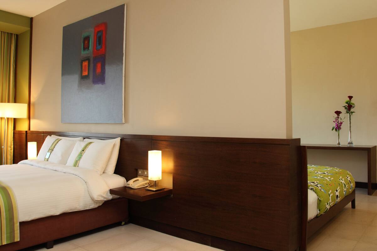 A hotel room with a bed, bedside lamps, a telephone, a painting on the wall, and a small table with flowers.