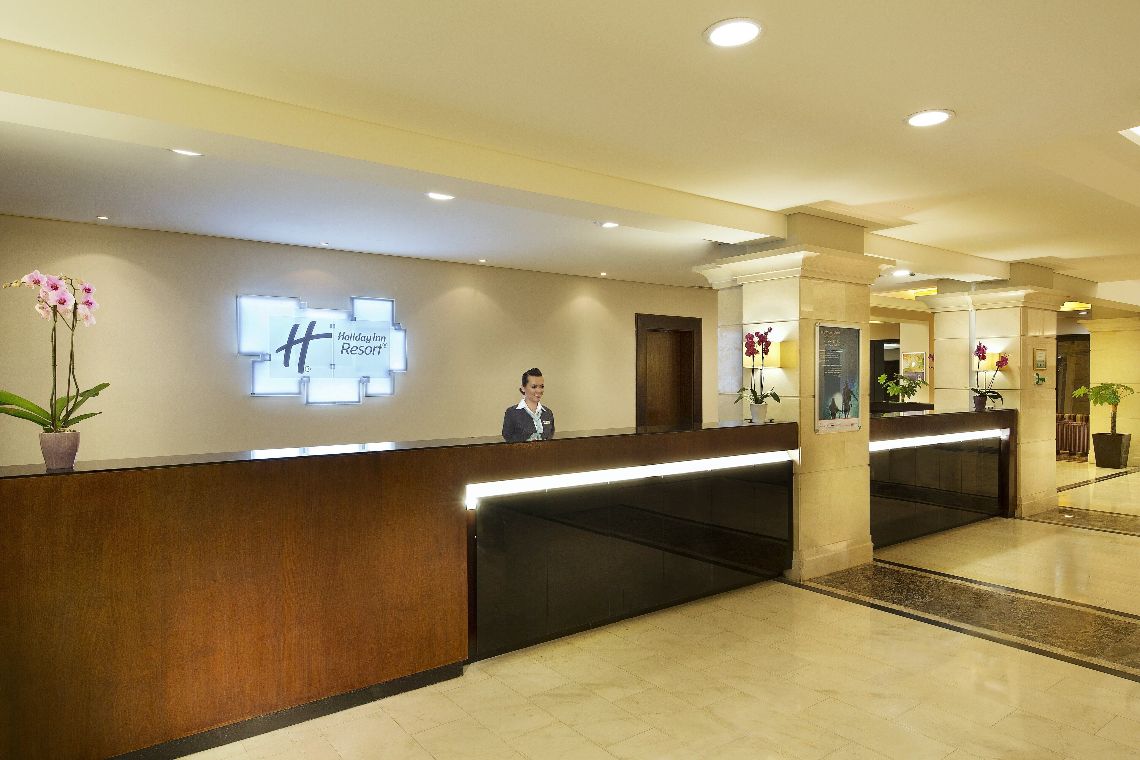 A hotel reception desk with a wooden counter, a digital display, and potted plants.