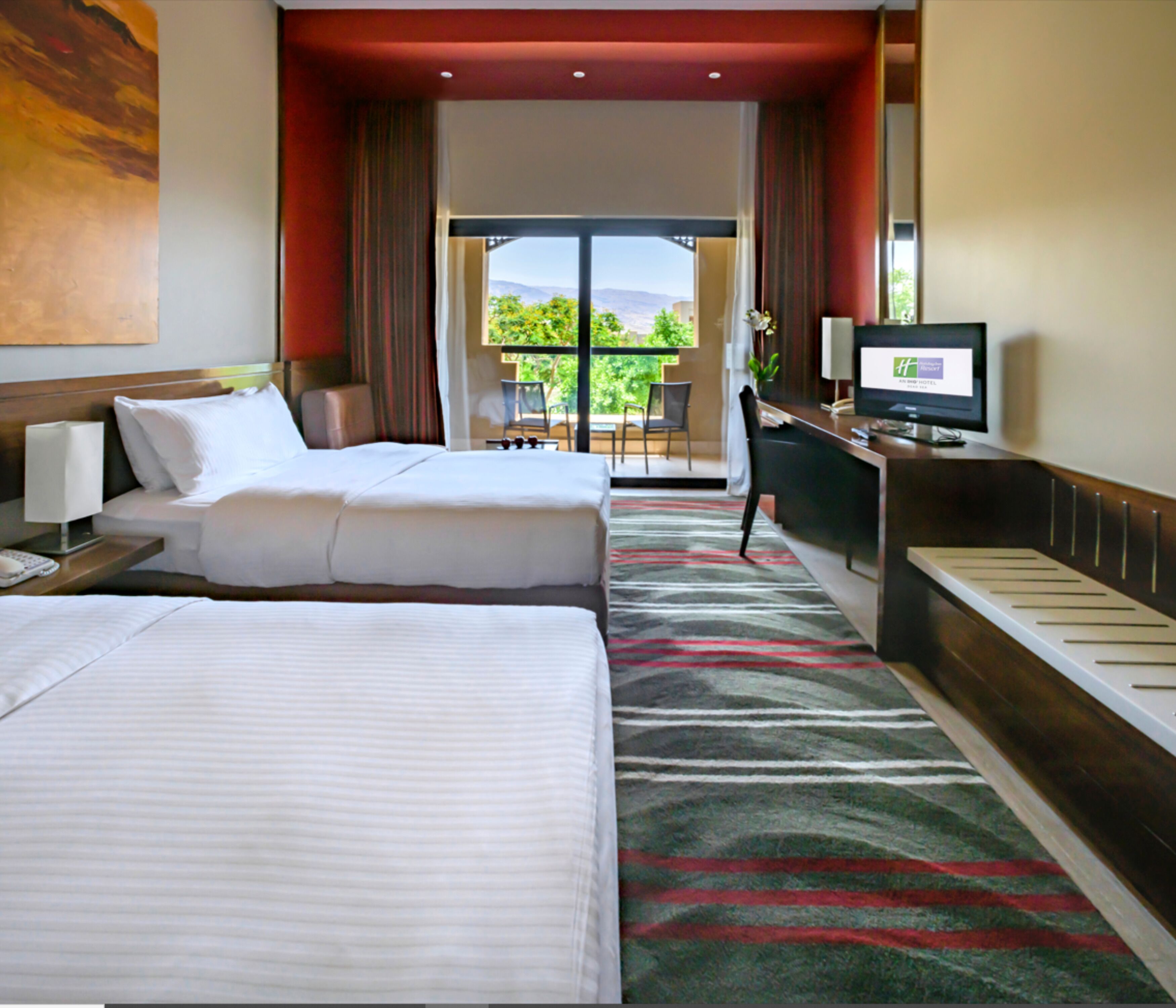A hotel room with two beds, a desk with a computer, a TV, and a view of the outdoors.