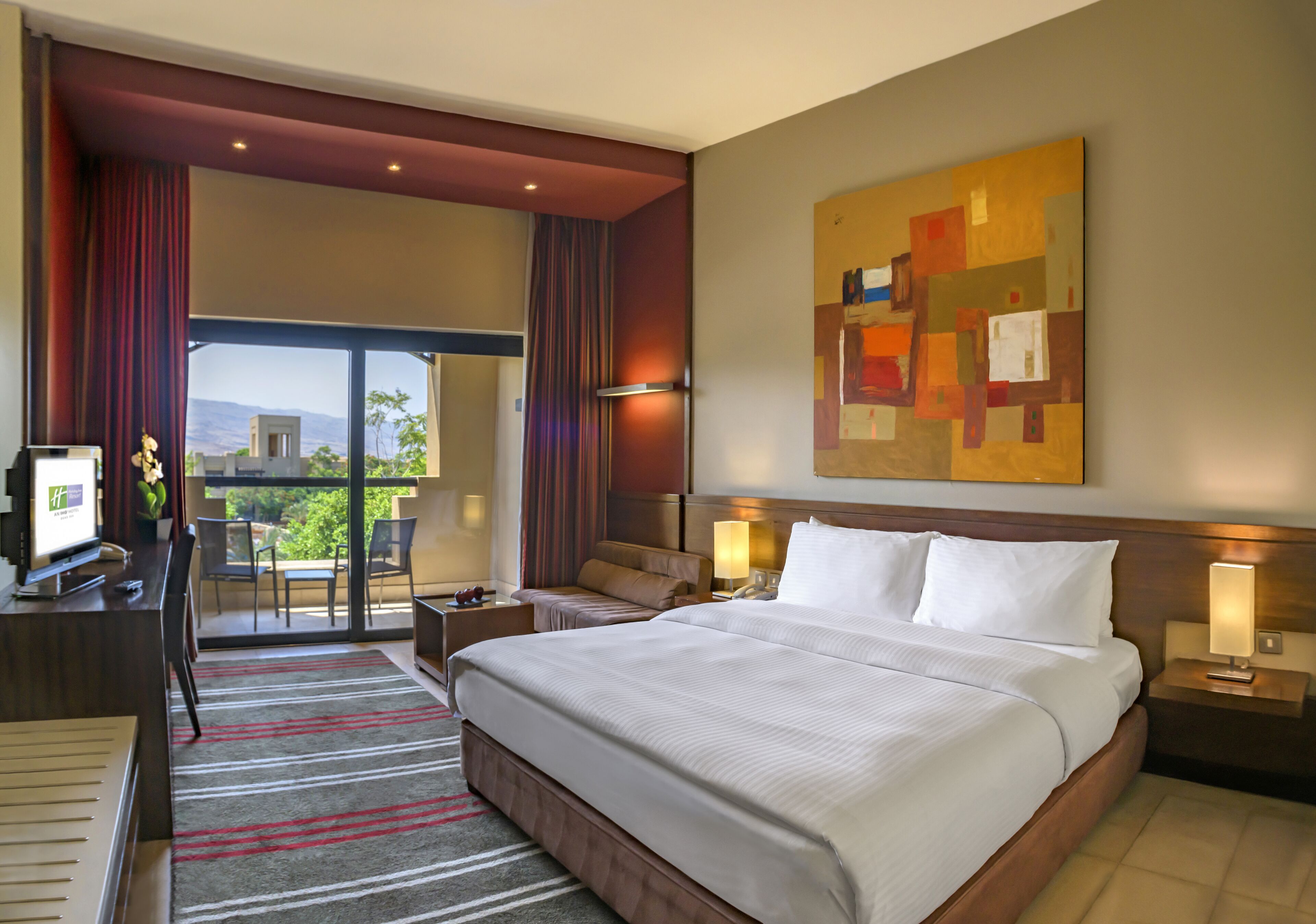 Une chambre d’hôtel avec un grand lit, un bureau avec un ordinateur, une chaise, une petite table, une lampe et un tableau au mur.