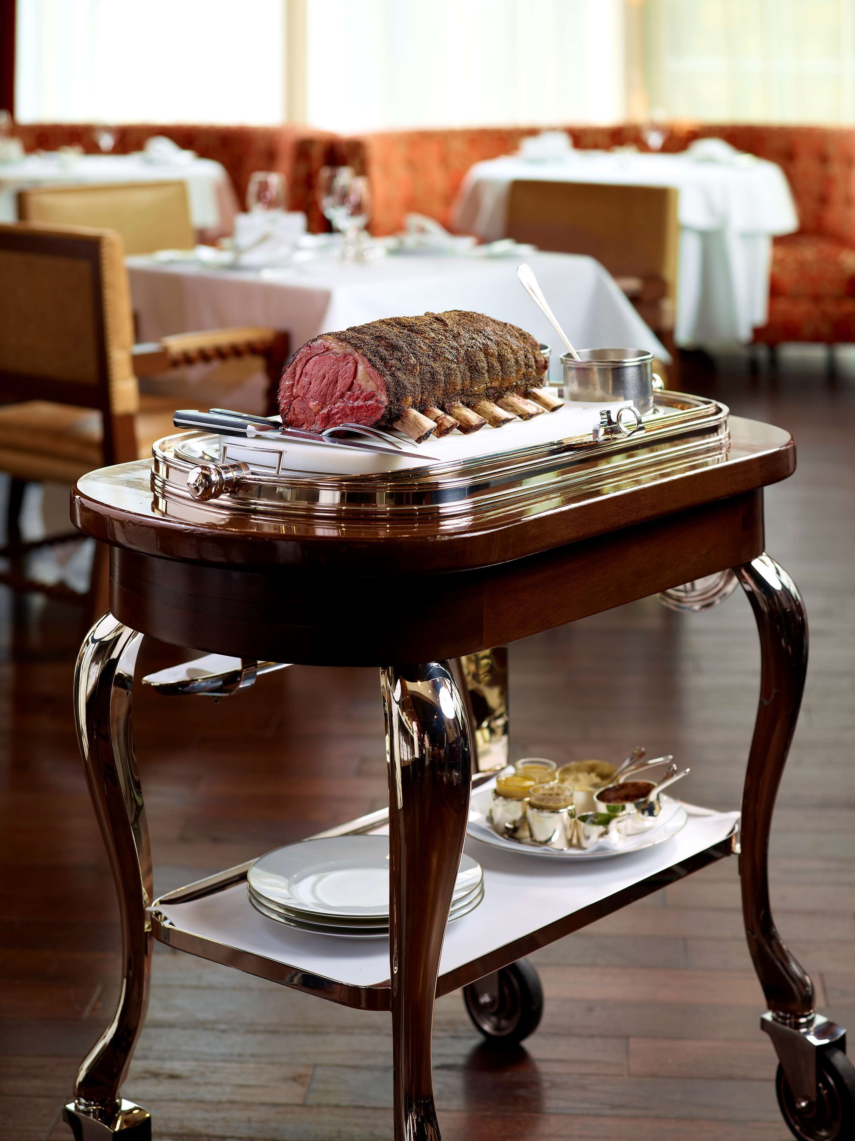 Una mesa de comedor con un plato de carne asada como centro, una bandeja con fuentes pequeñas y un carrito con platos.