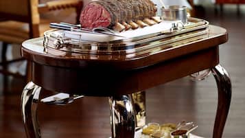 Una mesa de comedor con un plato de carne asada como centro, una bandeja con fuentes pequeñas y un carrito con platos.