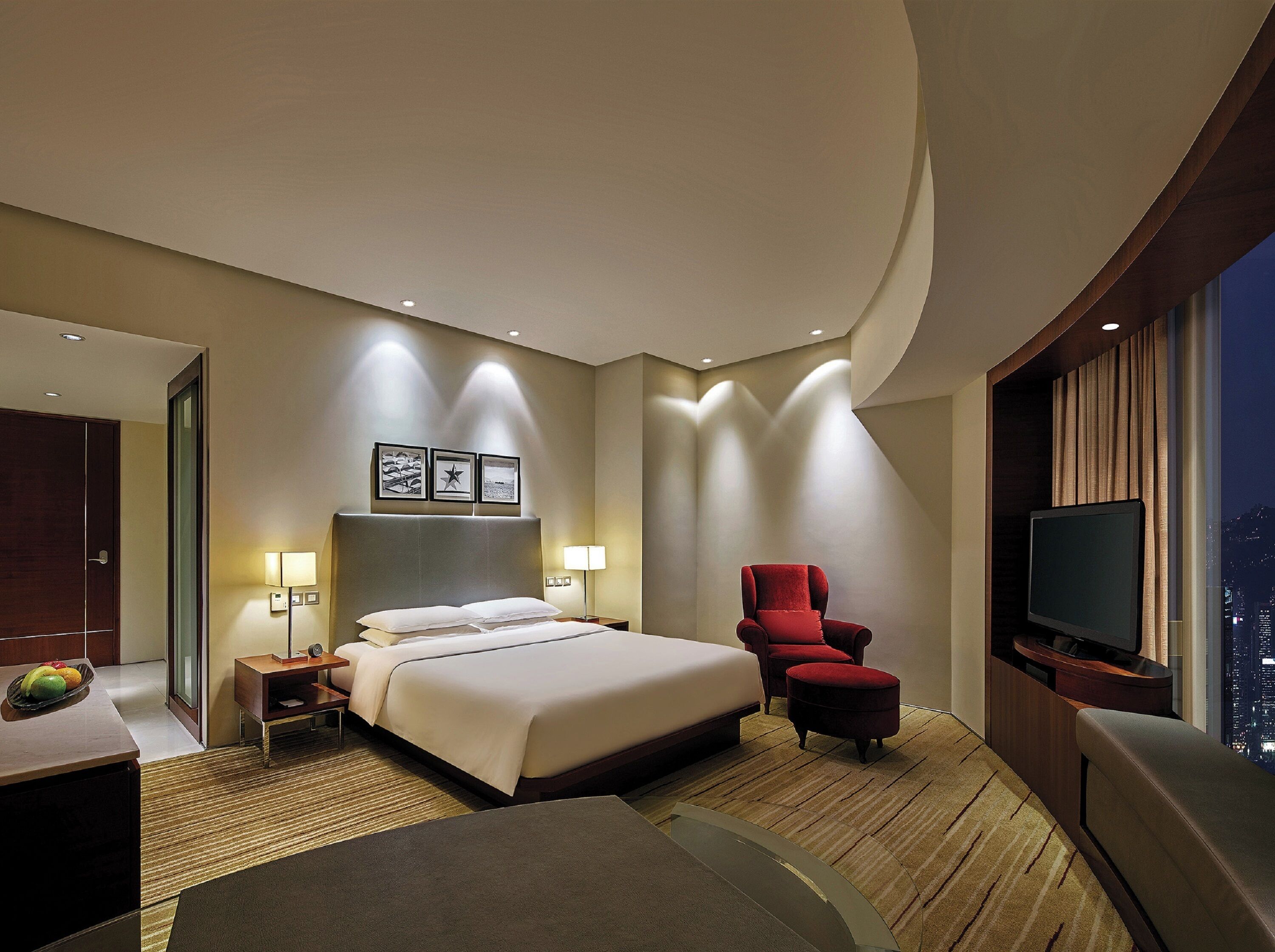 A modern hotel room with a large bed, a red armchair, a flat-screen TV, and a city view.