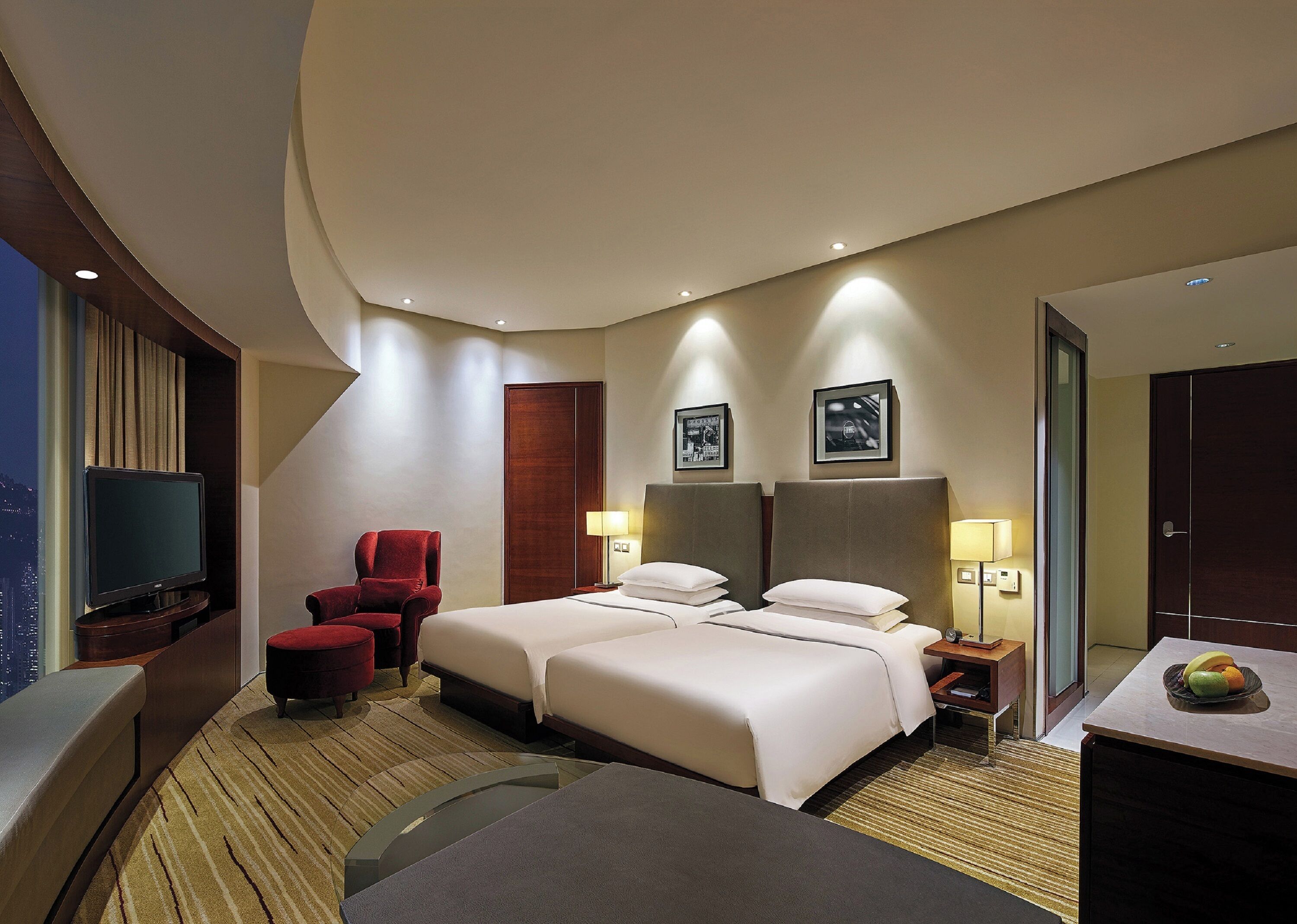 A hotel room with a large bed, a TV, a red chair, and a desk with a fruit bowl.