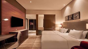 A hotel room with a bed, desk, chair, TV, and a red armchair.