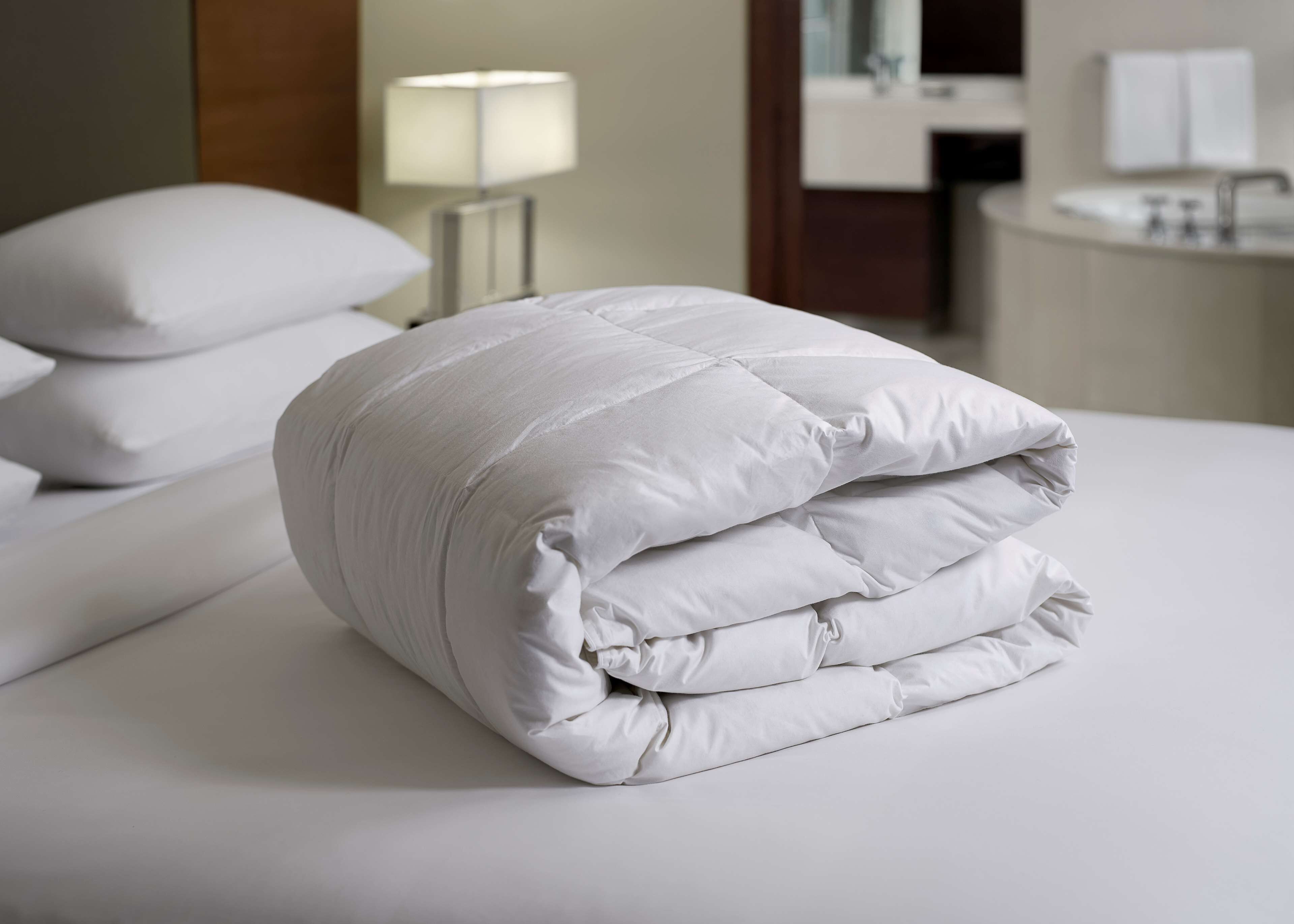 A neatly made bed with a white duvet and pillows, a bedside lamp, and a bathroom in the background.