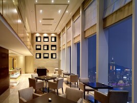 A modern hotel lobby with a city view, featuring a reception desk, seating area, and a wall with recessed lighting.