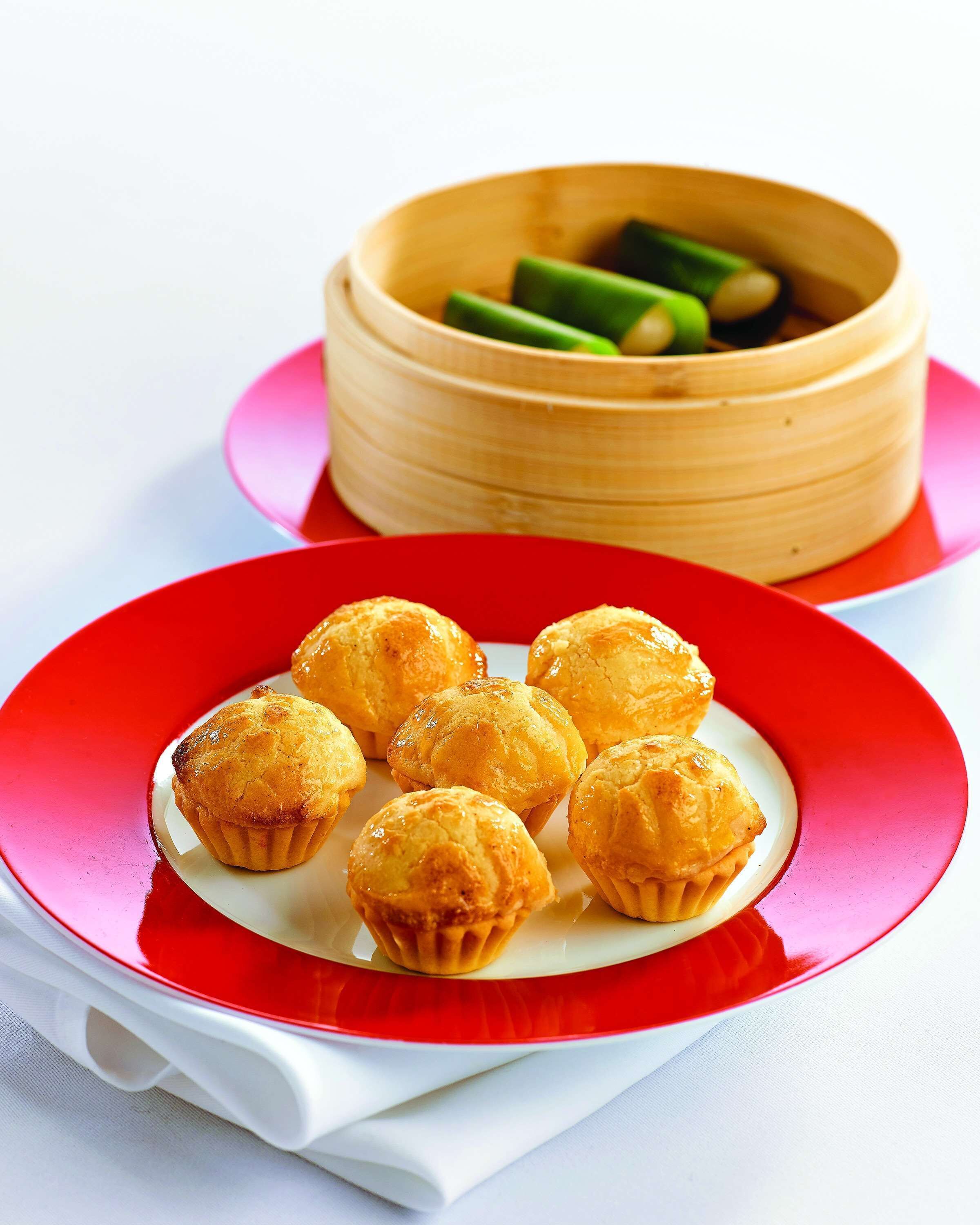 A plate of golden-brown pastries on a red plate, with a bamboo steamer in the background.