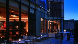 An outdoor seating area with tables and chairs, a bar area visible through the glass doors, and a building with a modern architectural design. - Hyatt Regency Hong Kong, Tsim Sha Tsui (Kowloon)