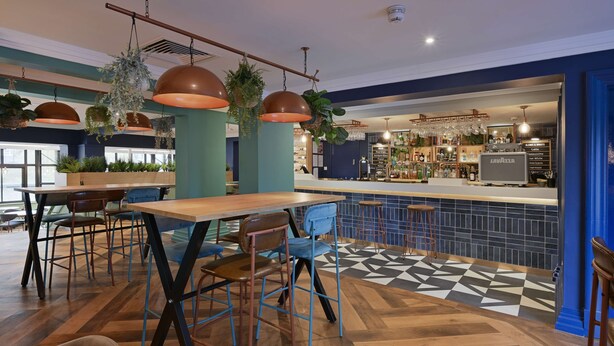 A modern bar area with wooden tables and blue chairs, a patterned floor, and a bar counter with a liquor display. - Travelodge Bath Waterside (Bath)