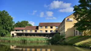 A hotel with a reflection in a calm body of water, featuring a covered outdoor area and a brick facade. - Travelodge Bath Waterside (Bath)