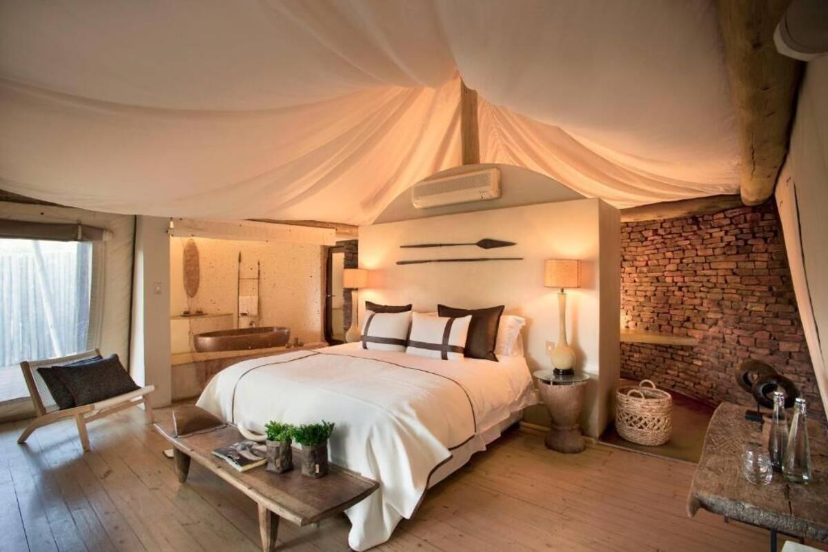 A bedroom with a large bed, a wooden bench, a bedside table with a lamp, a brick wall, and a window with curtains.