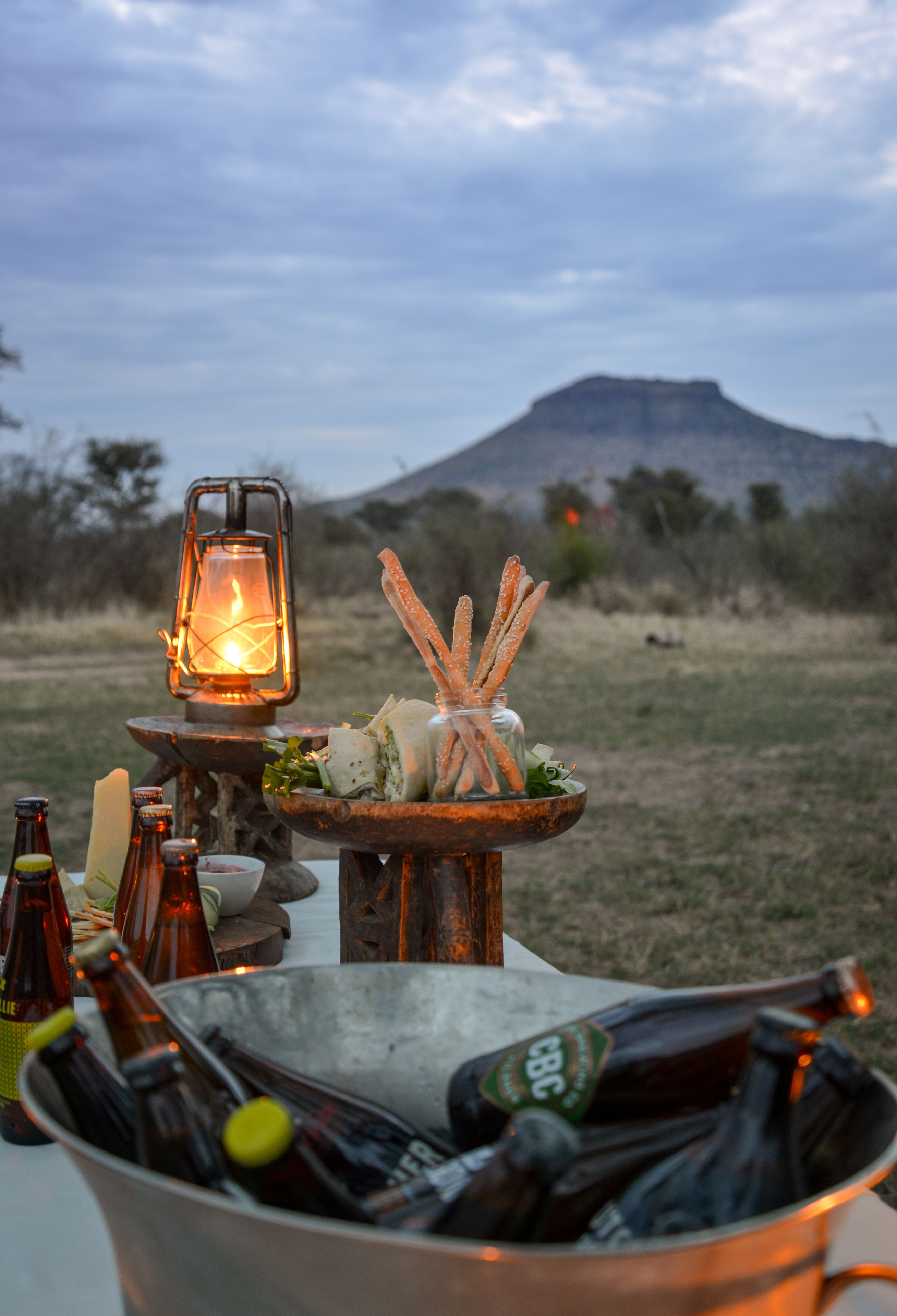 A rustic outdoor dining setup with a lantern, breadsticks, and a metal bucket filled with bottles.