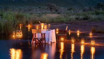 A dining setup with a table and chairs by a body of water, with a mountain in the background.