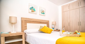 Un dormitorio con cabecera de madera, dos mesitas de noche, cama con ropa blanca y almohada amarilla, una mesita con lámpara, estante en la pared y armario.