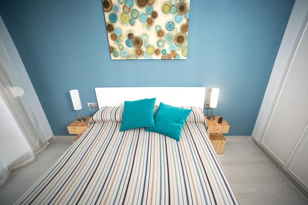 A bedroom with a striped bedspread, two teal pillows, a white headboard, and a wall art piece above the bed.