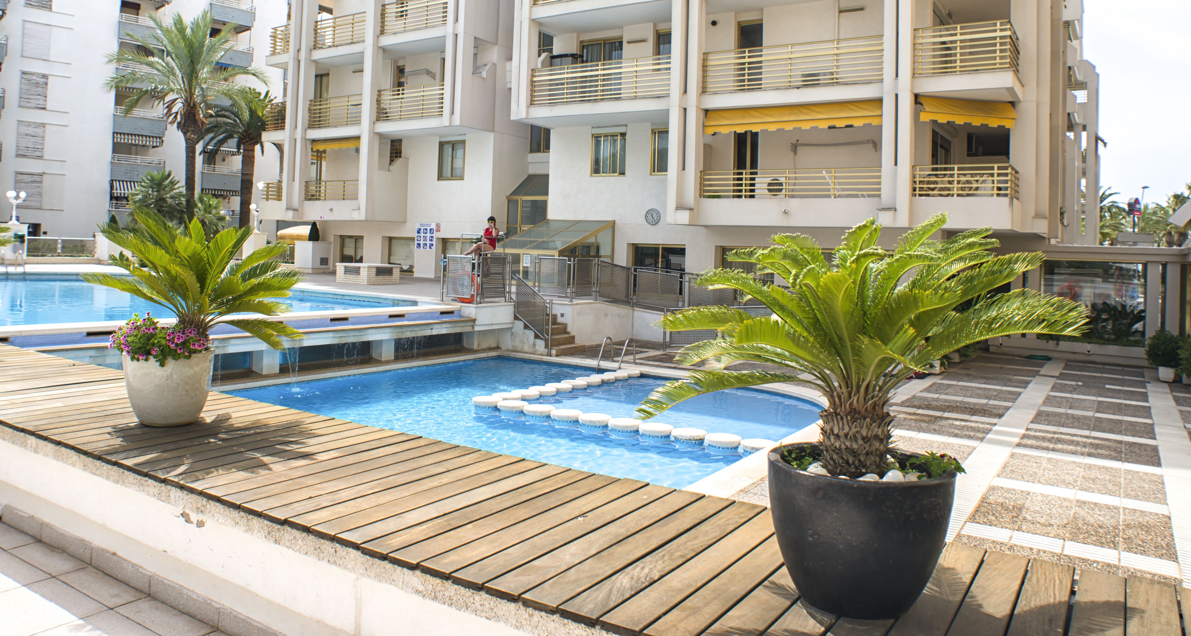 A rooftop pool area with a wooden deck, two potted plants, and a building with multiple balconies in the background.