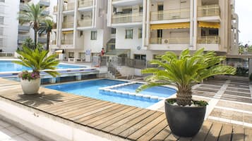 A rooftop pool area with a wooden deck, two potted plants, and a building with multiple balconies in the background.