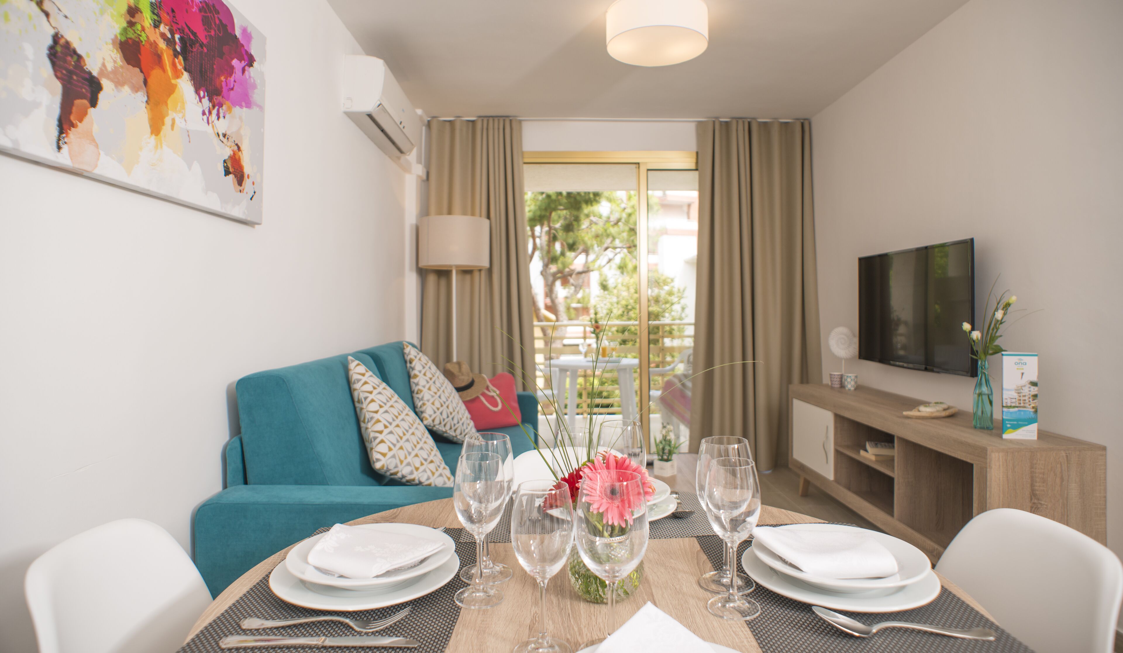 Un coin repas avec une table dressée pour quatre personnes, un canapé turquoise agrémenté de coussins à motifs, un téléviseur fixé au mur et un balcon avec des rideaux.