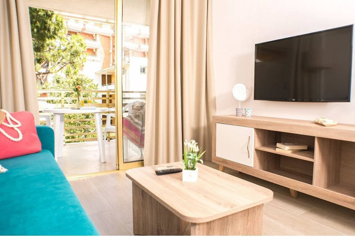 A modern living room with a teal sofa, a wooden coffee table, a TV mounted on the wall, and a view of the outdoor balcony.