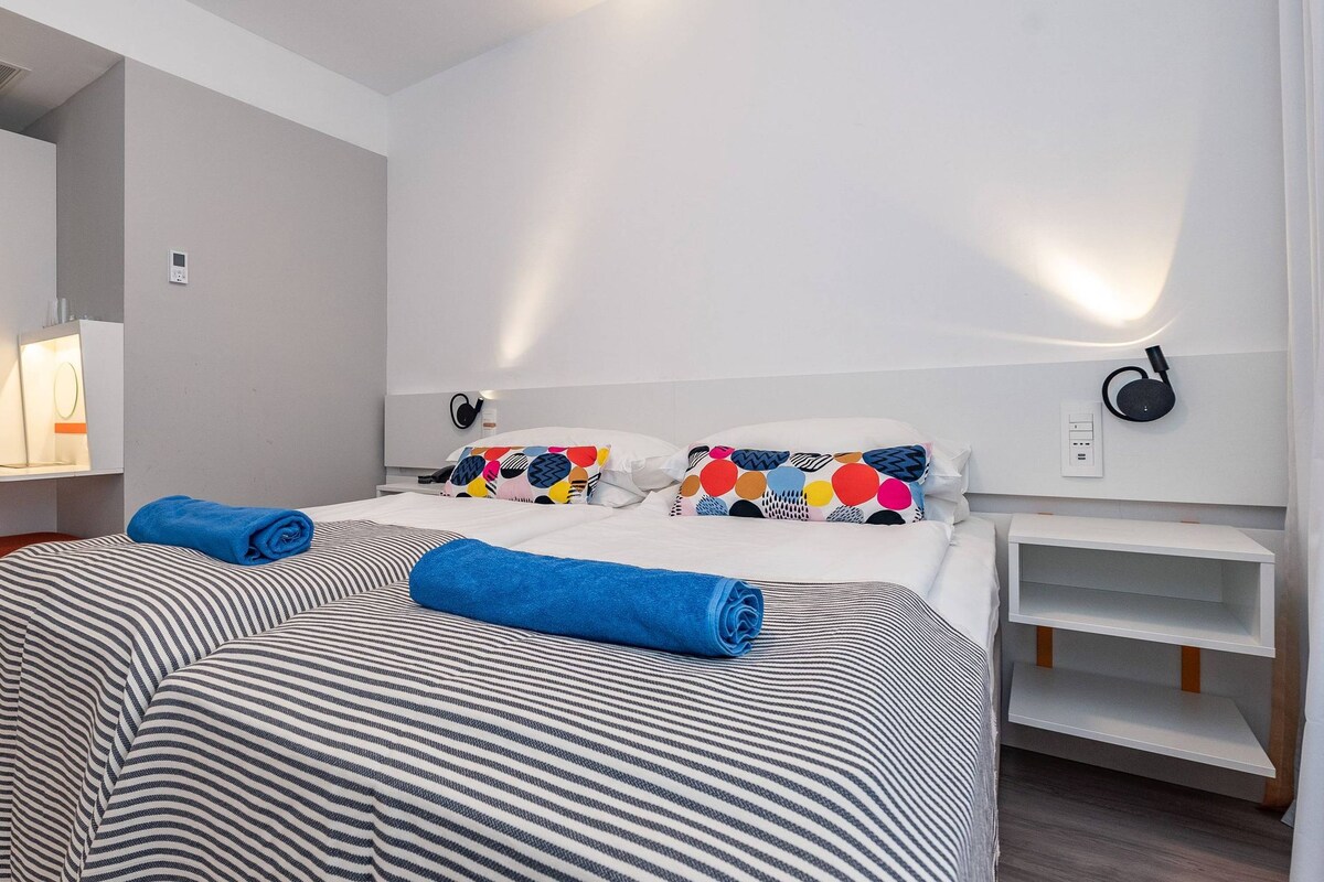 A hotel room with a bed, a bedside table, a lamp, a wall-mounted speaker, and a wardrobe.