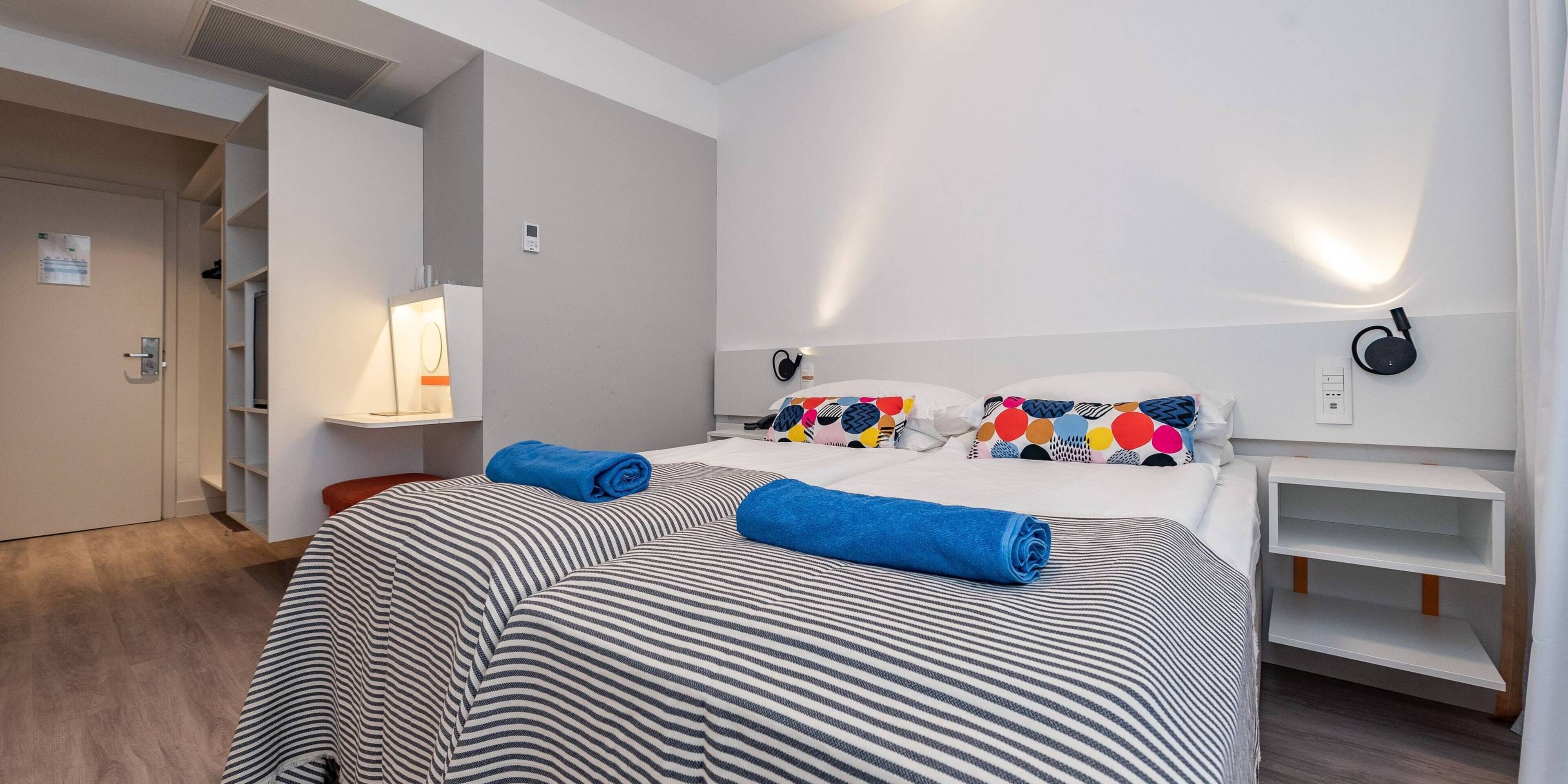 Quarto de hotel com cama, mesa de cabeceira, luminária, alto-falante fixado na parede e guarda-roupa.