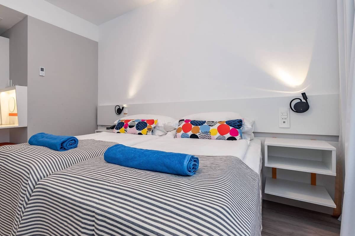 Una habitación de hotel con cama, mesita de noche, lámpara, altavoz en la pared y armario.