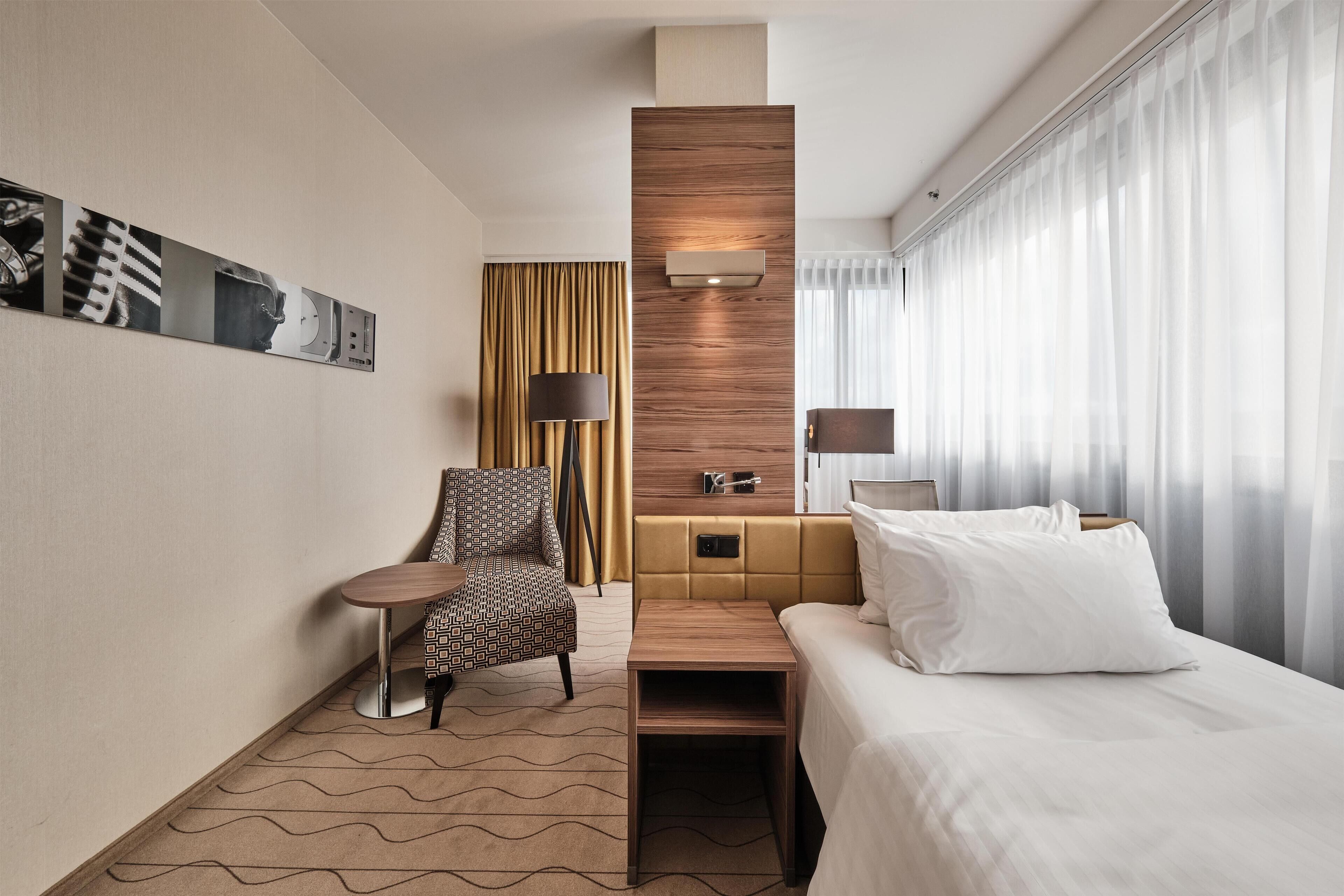 A hotel room with a bed, a chair, a small table, a sofa, a wall-mounted lamp, a wooden accent wall, and a window with curtains.