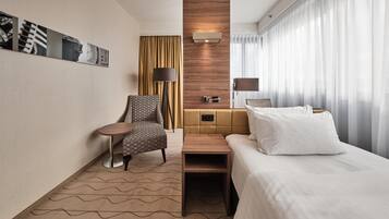 A hotel room with a bed, a chair, a small table, a sofa, a wall-mounted lamp, a wooden accent wall, and a window with curtains.