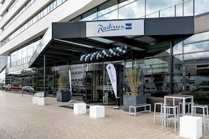 A Radisson BLU hotel with a modern glass facade, outdoor seating area, and a clear sky.