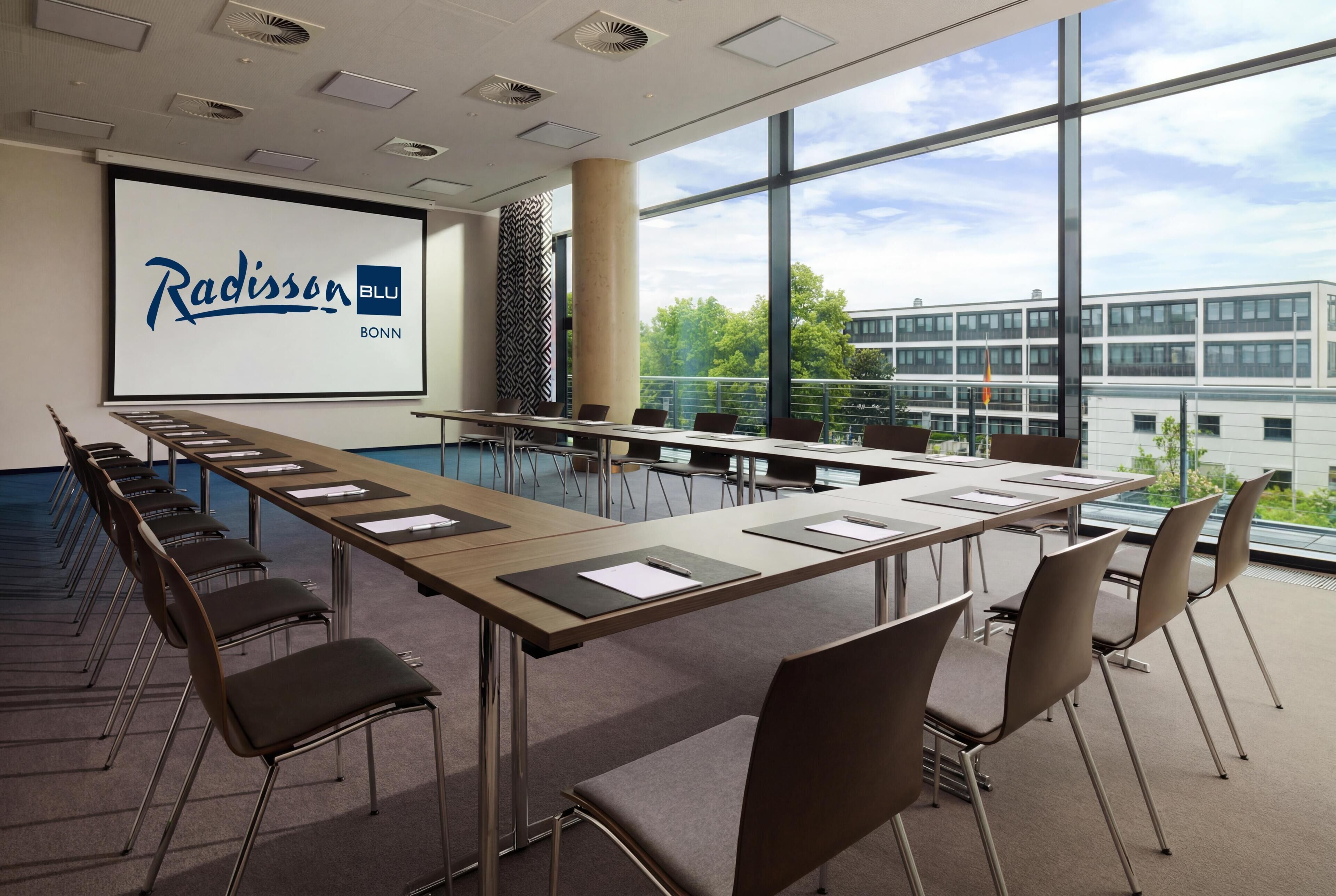 A conference room with a large screen displaying Radisson BLU Bonn, a long table with chairs, and large windows offering a view of buildings and trees.