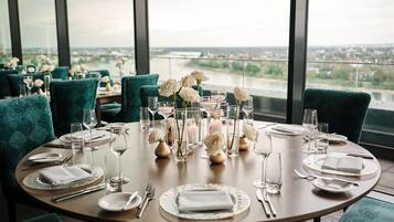 A round dining table with a view of a river and cityscape, set with white roses and candles.
